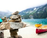 Steinbalance am Ufer eines Sees mit bunten Kanus und bewaldeten Bergen im Hintergrund.